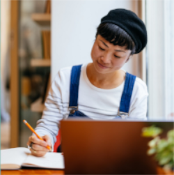 A picture of an Asian woman in a striped shirt and overalls sitting in
			front of a laptop with a pencil in her hand.